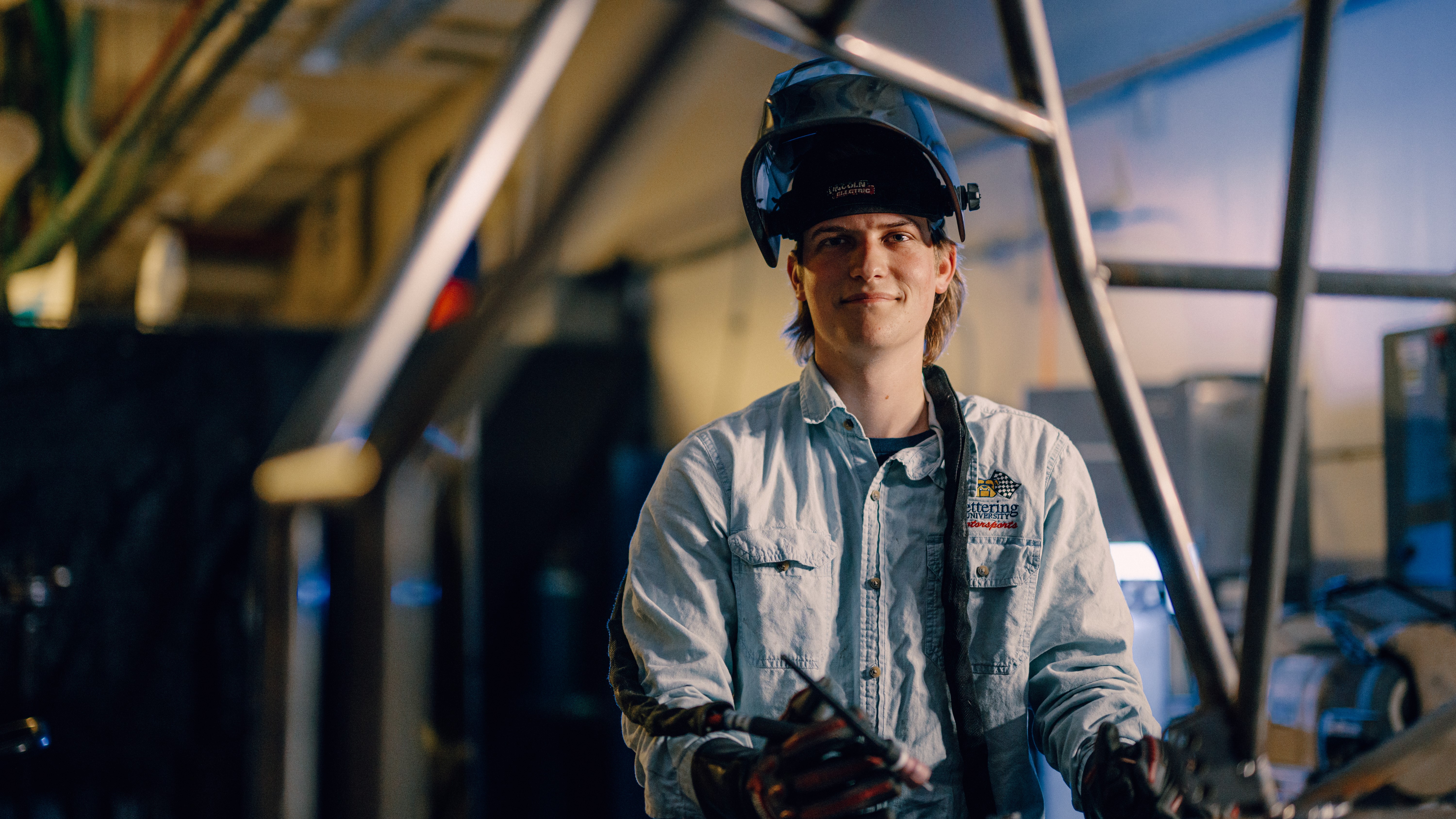 A Kettering student wears a helmet and racing goves and holds a tool. He stands in the SAE Garage.
