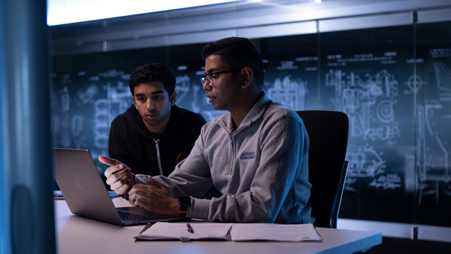 Two students sit at a table looking at a laptop