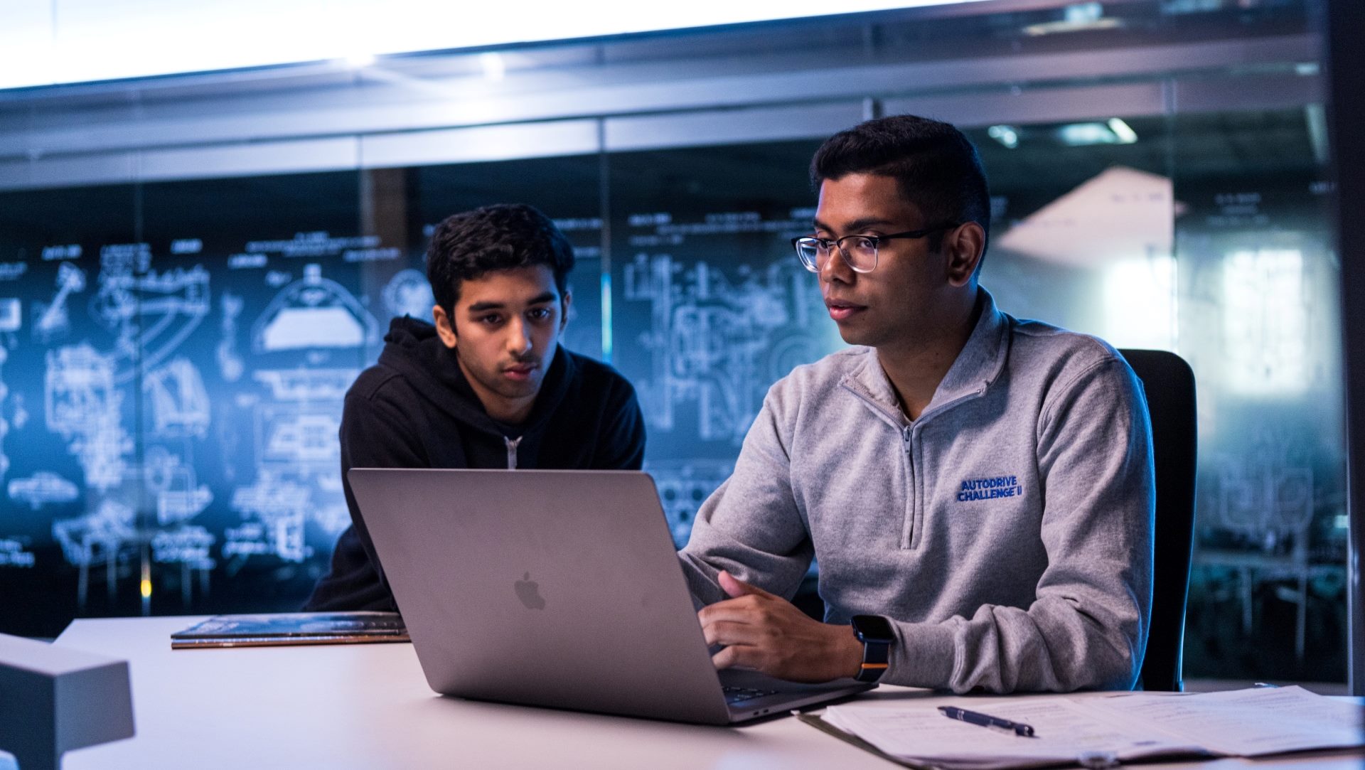 Two students sit at a table looking at a laptop