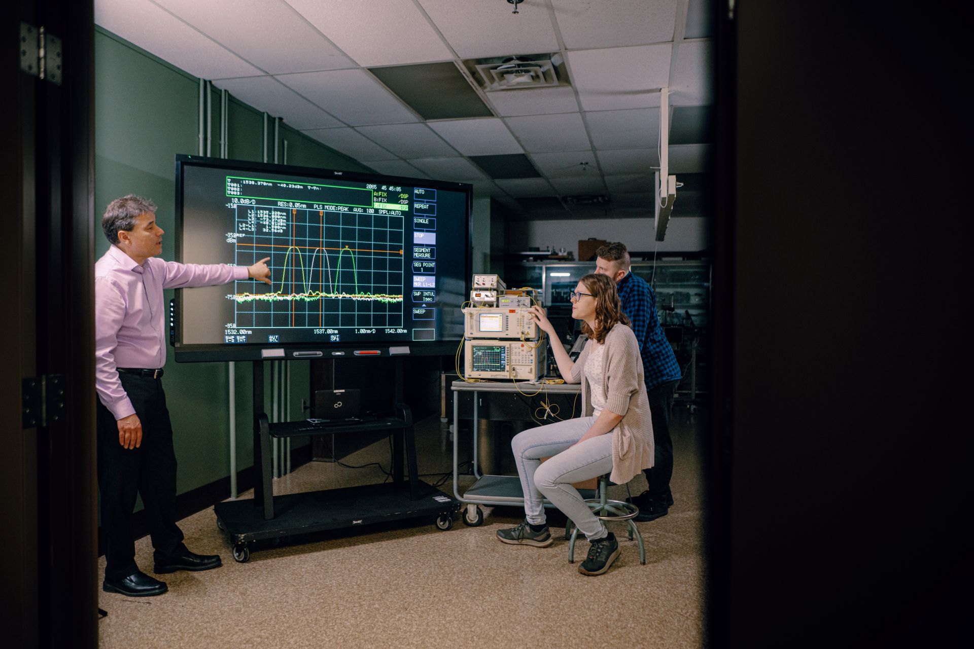 Data Science professor and two students analyzing signal output on a large screen with optics lab computer equipment nearby