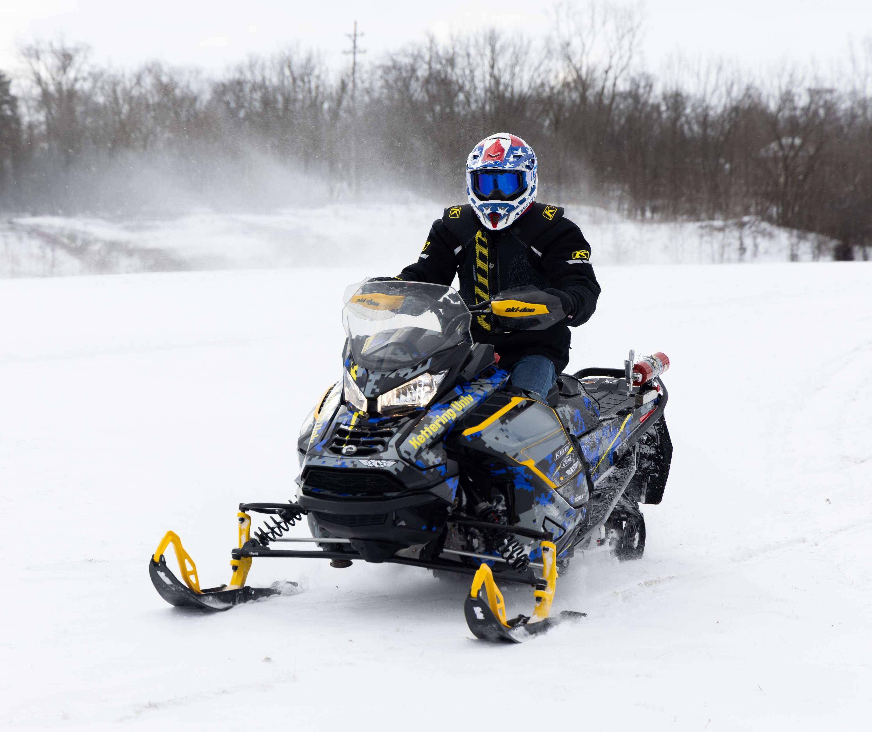 Student rides a snowmobile across a snow-covered field, wearing a full-face helmet and winter gear, as snow kicks up behind the machine; the vehicle features Kettering University branding in blue and yellow.