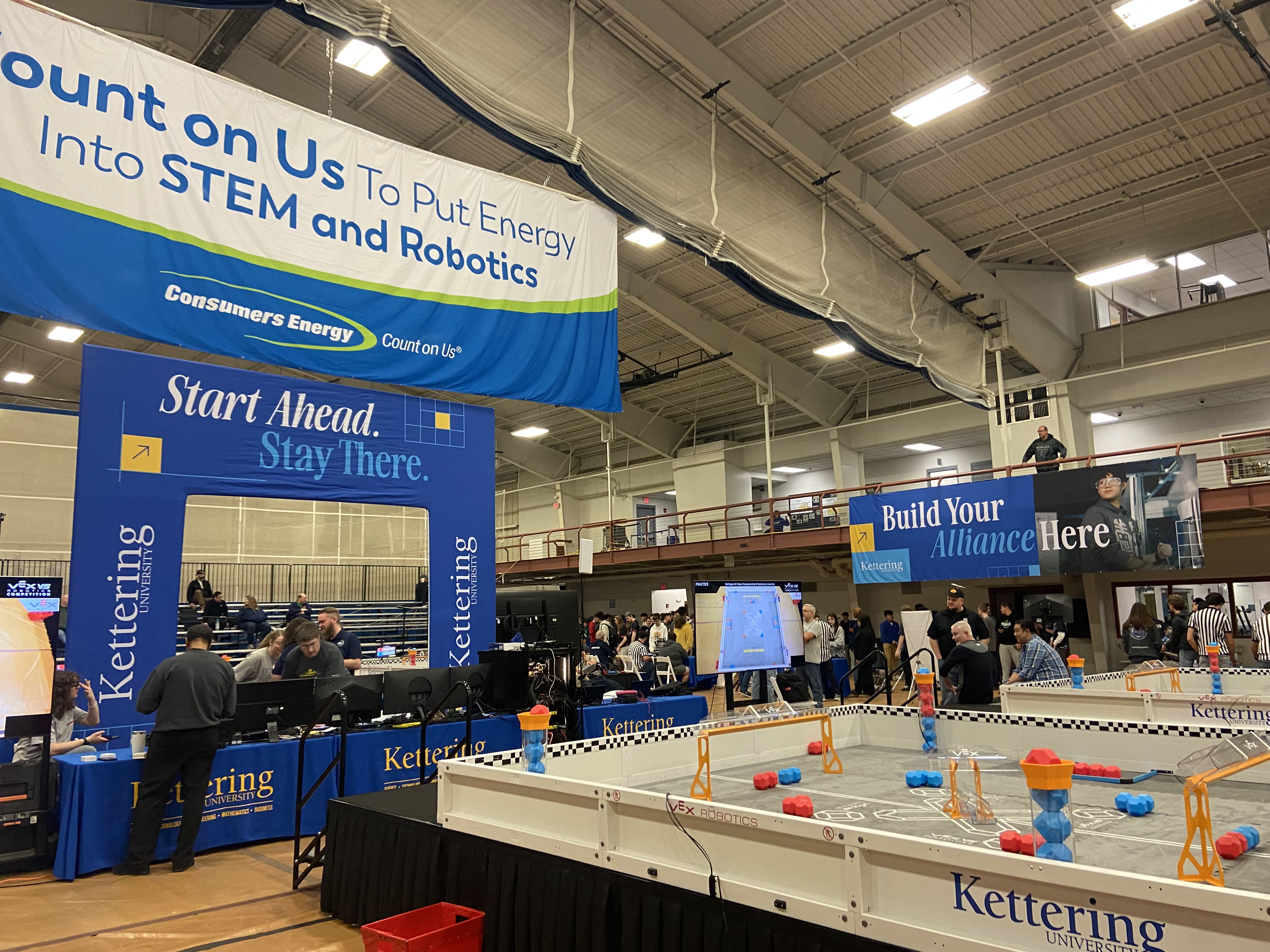 A view of the Connie and Jim John Recreation Center at Kettering University during the  VEX Robotics State Championship. Consumers engergy banners hang from the ceiling and multiple robotics event fields are active with competition. 