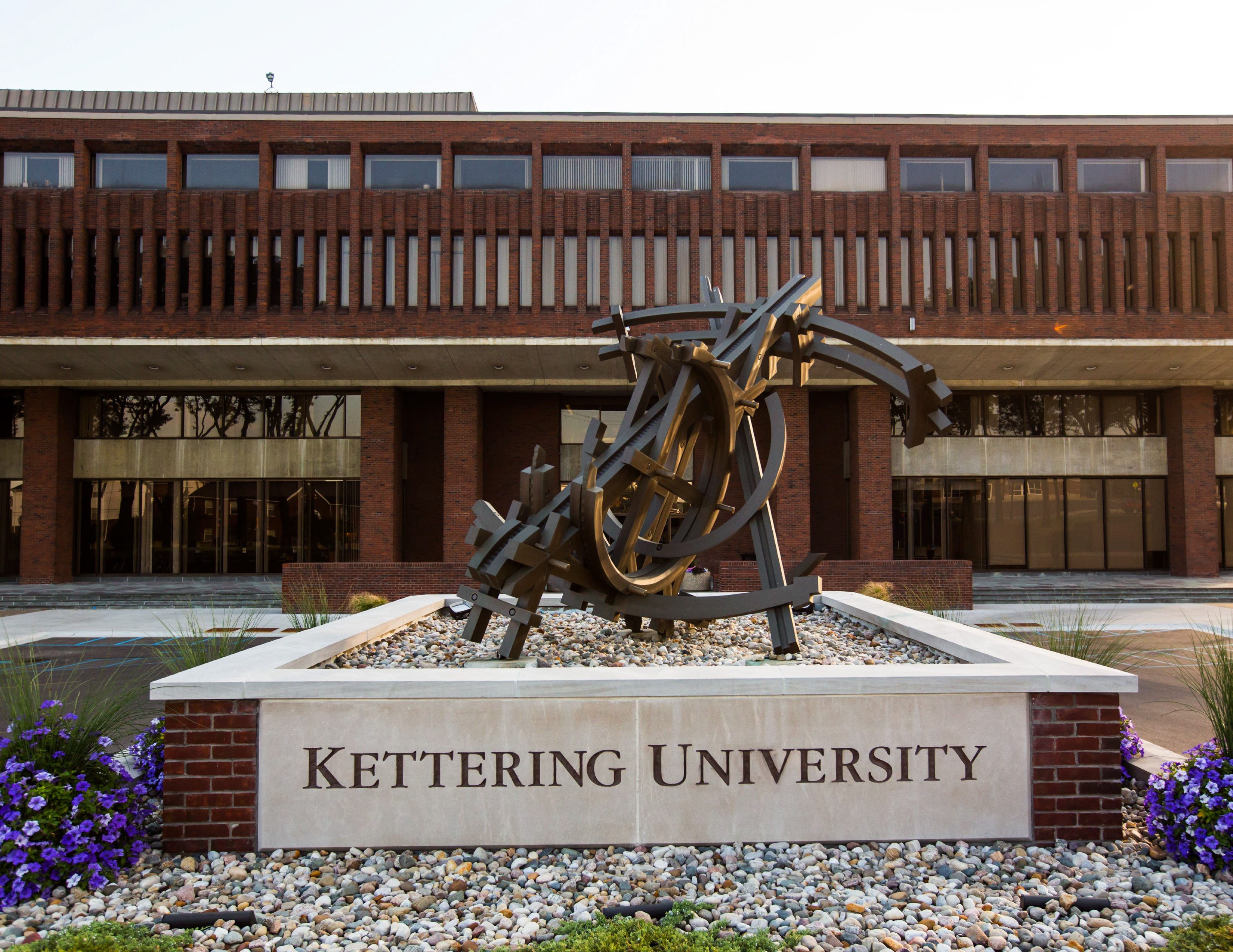 Exterior view of Kettering University’s main campus entrance featuring the university name engraved on a stone monument beneath a large abstract metal sculpture, with the brick academic building and glass entryways in the background.
