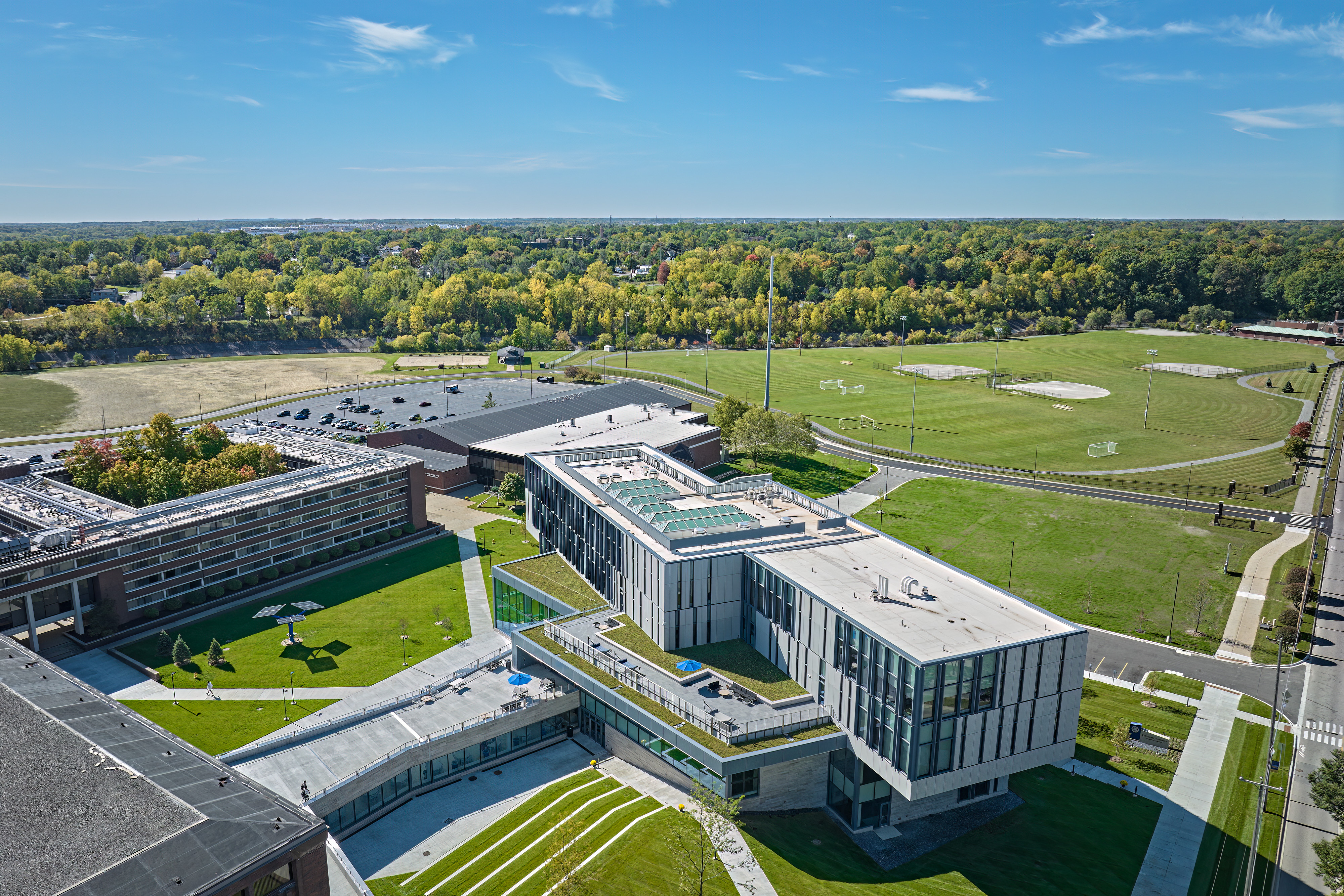 Aerial view of Kettering University's Learning Commons on a sunny autumn day. The modern academic building features expansive glass walls, rooftop terraces, and green roofs, with landscaped walkways connecting to surrounding campus buildings. Athletic fields, parking areas, and tree-lined neighborhoods extend into the distance beneath a clear blue sky.