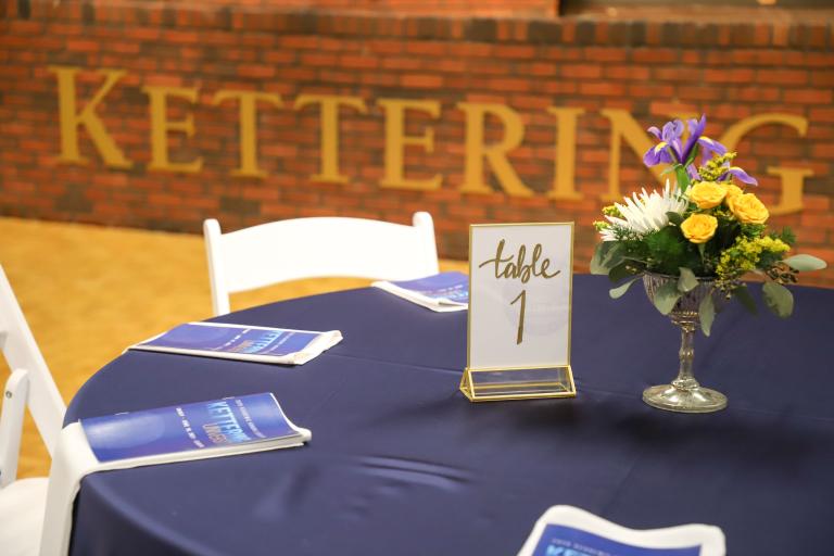 Programs, a table number card flower centerpiece sit on a table covered with a blue tablecloth