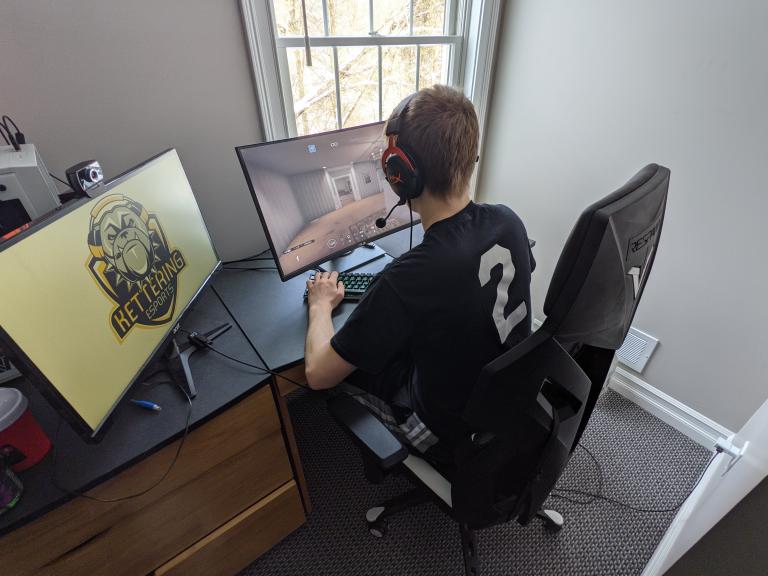 A male Kettering University student plays Rainbow Siege Six on the right screen while the Kettering University Esports logo is displayed on the left screen.