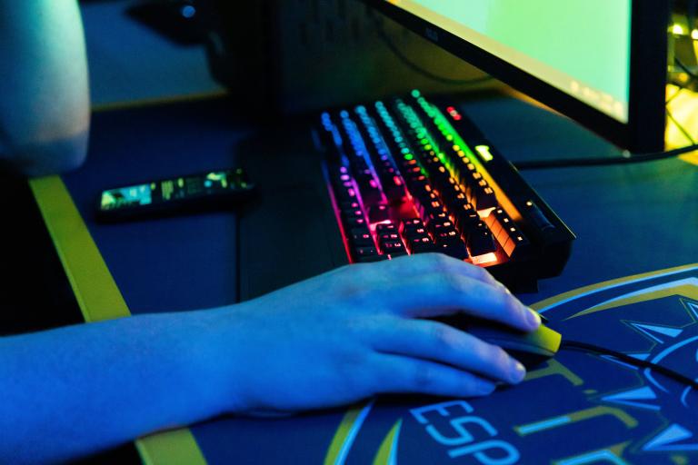 A hand on a mouse with a keyboard illuminated with rainbow lights.