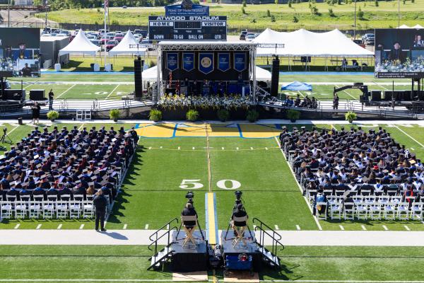 Wide view of Kettering University's 2026 Commencement Ceremony at Atwood Stadium, with graduates seated on the field facing the commencement stage and scoreboard.