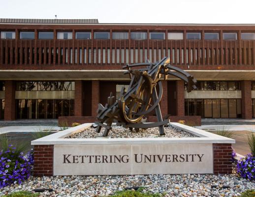 Exterior view of Kettering University’s main campus entrance featuring the university name engraved on a stone monument beneath a large abstract metal sculpture, with the brick academic building and glass entryways in the background.