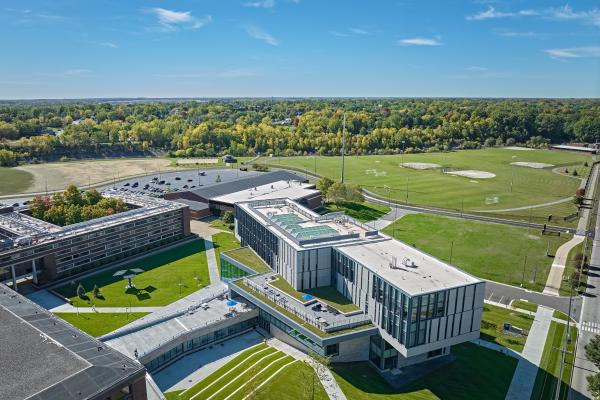 Aerial view of Kettering University's Learning Commons on a sunny autumn day. The modern academic building features expansive glass walls, rooftop terraces, and green roofs, with landscaped walkways connecting to surrounding campus buildings. Athletic fields, parking areas, and tree-lined neighborhoods extend into the distance beneath a clear blue sky.