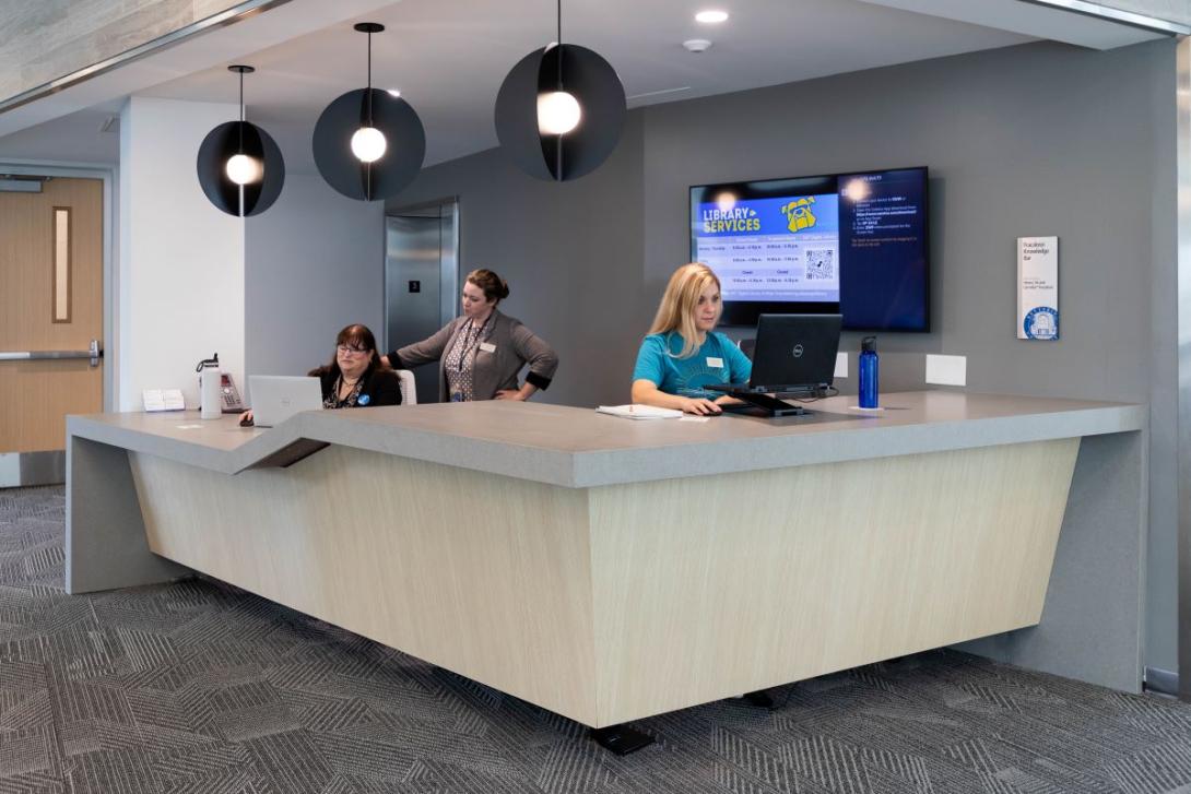 Two Kettering employees and a student worker work at computers at the Library Services desk in the Learning Commons